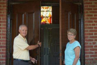 Deacon Greg Beavin and his wife Shirley stand at the entrance of St. Theresa Church in Rhodelia, Ky., Aug. 3. St. Theresa is the oldest church in Meade County and it’s one of the parishes where Deacon Beavin was assigned after ordination. The deacon and his wife grew up at the parish. (Record Photo by Ruby Thomas)